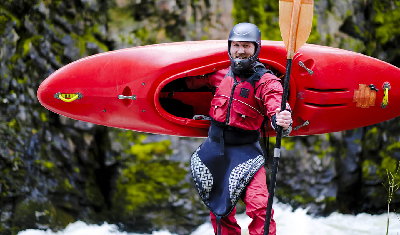 Man wearing a kayak spray skirt carrying his red kayak and a paddle